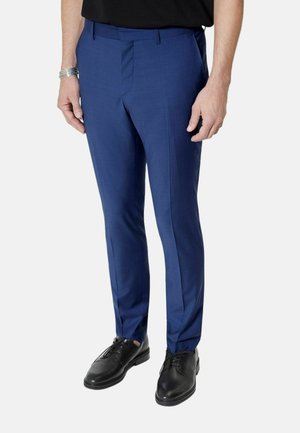 Mand iført smalle blå habitbukser, sorte lædersko og sort skjorte, stående foran en ensfarvet lys baggrund.