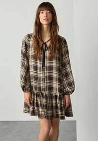 Robe à carreaux noire et beige avec un ourlet froncé, des manches longues bouffantes et un détail à nouer au col. Fabriquée en tissu léger.