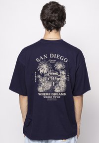 Marineblaue Baumwoll-T-Shirt mit einem großen beigen Grafik, die ein Gebäude und Palmen zeigt, mit dem Text "San Diego" und "Kaotiko Resort."