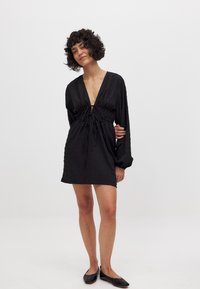 Čierne mini šaty s dlhými, objemnými rukávmi, hlbokým výstrihom do V a naťahovacím pásom. Textúrovaná látka s voľným strihom a detailom na stiahnutie.