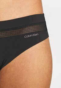 Black bikini bottom with a smooth texture, featuring a mesh accent on the waistband and a printed "Calvin Klein" logo.
