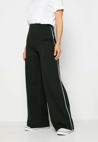 Pantalons larges noirs en tissu doux. Présentent des rayures latérales blanches et une taille haute. Assorties à des baskets blanches.