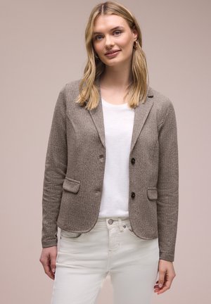 Jonge vrouw met schouderlang blond haar, gekleed in een grijze textuurblazer, wit T-shirt en witte broek, staand tegen een neutrale achtergrond.