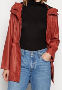 Rød, letvægts, vandtæt jakke med hætte, frontlynlås, trykknapper og elastisk talje. Bæres over en sort højhalset trøje og blå jeans.
