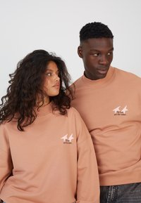 Two models wear matching oversized, peach-colored sweatshirts. Each features a white printed graphic with the text "No two are the same."