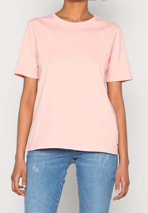 Kobieta w jasnoróżowej koszulce Tommy Hilfiger w połączeniu z niebieskimi przetartymi dżinsami, stojąca z rękami swobodnie opartymi po bokach.