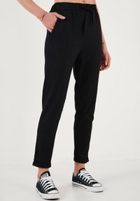 Zwarte tapse broek van een textiele stof, met een elastische tailleband en een externe trekkoord; zijzakken; gedragen met zwart-witte sneakers.