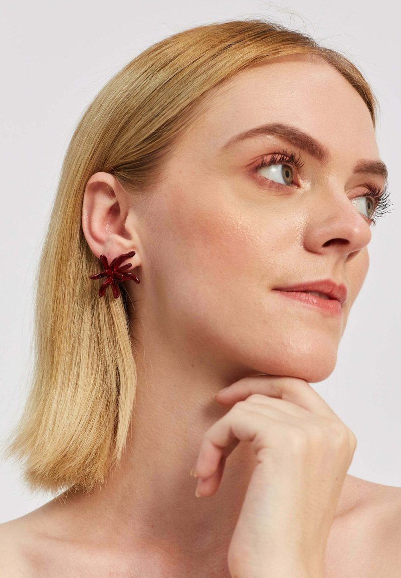 Červené akrylové náušnice v tvare hviezdy s ostrými špicami, s lesklým povrchom a žiarivým vzhľadom, ktoré nosí osoba s blond vlasmi.