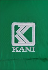 Grön tyg med en vit broderad logotyp som har ett rektangulärt mönster och en stiliserad 'K'-design, med texten "KANI" nedanför.