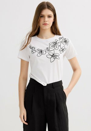 Joven mujer con cabello largo que lleva una camisa blanca de manga corta con arte floral en línea negra y pantalones negros de cintura alta, mano en el bolsillo.