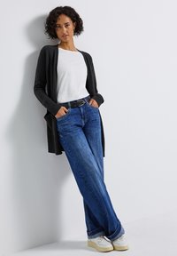 Grijze lange cardigan over een wit T-shirt, gecombineerd met lichtblauwe brede jeans en witte sneakers met gele zolen.