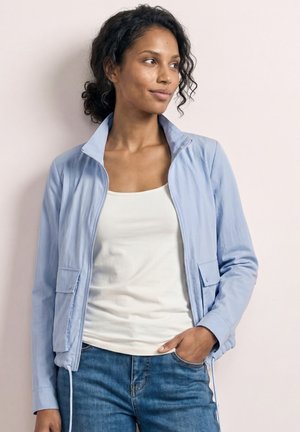 Kvinde med krøllet hår iført en lyseblå lynlåstrøje over en hvid tanktop og blå jeans, stående med den ene hånd i lommen og ser til siden.