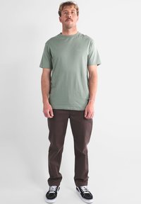 T-shirt en coton vert clair à manches courtes, associé à un pantalon marron foncé. Des baskets noires et blanches complètent la tenue.