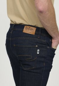 Jeans en denim bleu foncé avec une coupe fuselée, présentant des coutures orange, un patch en cuir sur l'arrière et des détails de poches accentués.