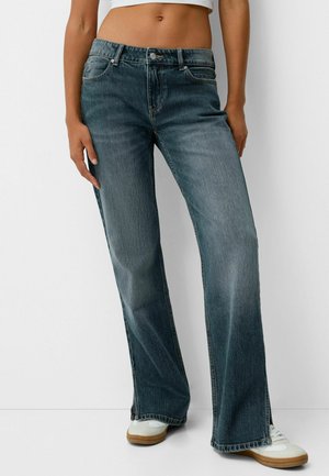 Vrouw draagt verweerde wijde blauwe jeans, witte sneakers en een wit cropped topje, staand tegen een effen witte achtergrond.