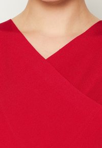 Červená látka s designem ve tvaru V, která má texturovaný, žebrovaný vzor. Překrývající se panely vytvářejí vrstevnatý vzhled v oblasti dekoltu.