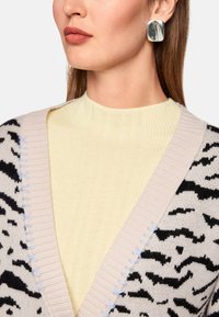 Gestrickte Strickjacke mit Animal-Print, beige und schwarz, mit blauen Nähten am Rand. Darunter ein geripptes, gelbes Oberteil mit hohem Kragen und silbernen quadratischen Ohrringen.