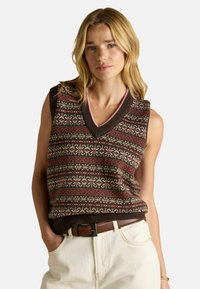 Blonde ženska, oblečena v vzorčen rjav brezrokavnik s klasičnim V-izrezom v roza odtenku, bež hlače in rjav pas, roke v žepih.