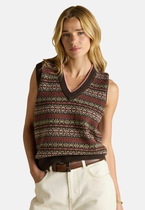 Blonde woman wearing a patterned brown sleeveless sweater vest with a pink-trimmed V-neck, beige pants, and a brown belt, hands in pockets.