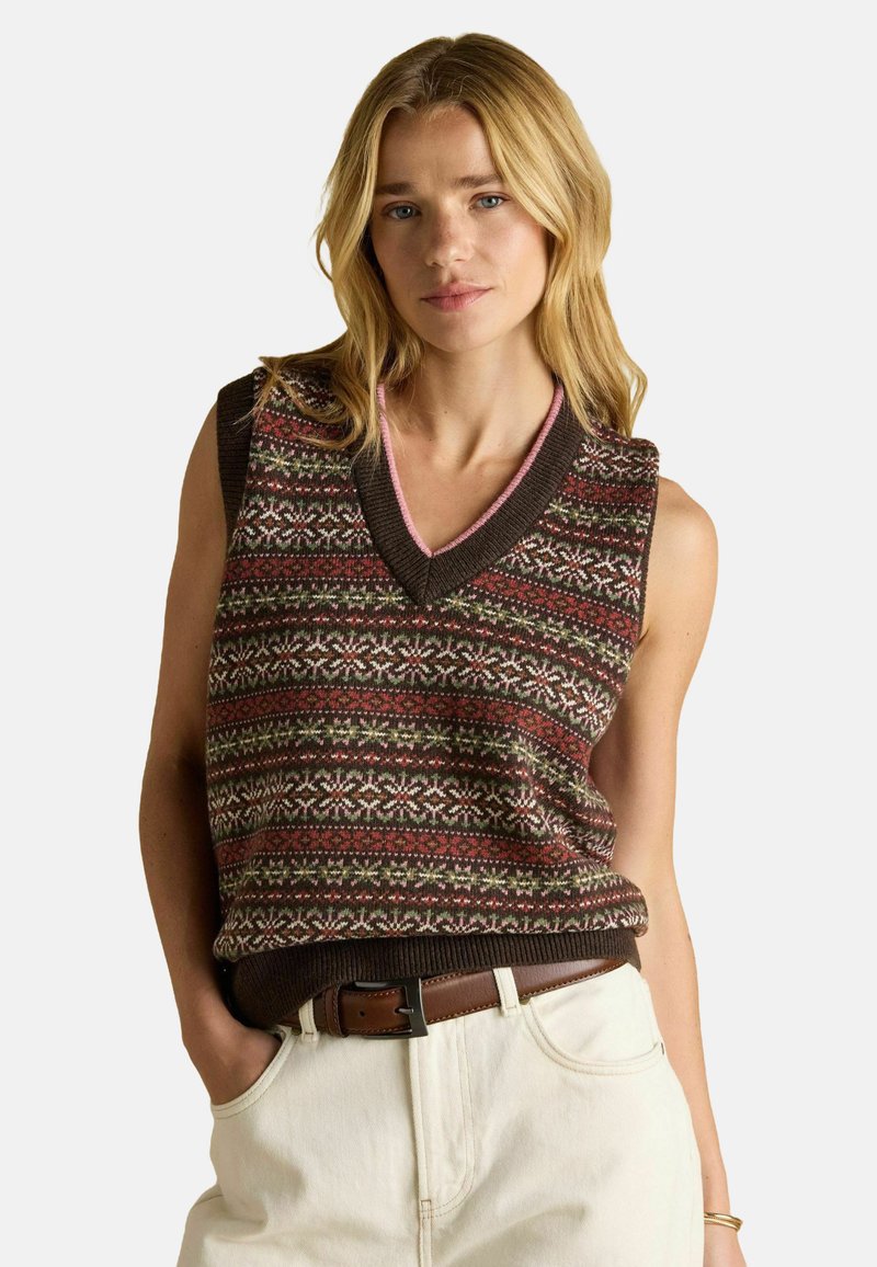 Blonde ženska, oblečena v vzorčen rjav brezrokavnik s klasičnim V-izrezom v roza odtenku, bež hlače in rjav pas, roke v žepih.