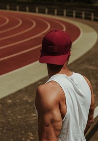 Maroon baseball cap with mesh panel, logo patch on front, worn by a person in a white tank top, standing on a red athletic track.