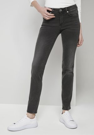 Slim-fit black jeans worn with white sneakers and a white long-sleeve top, model standing against a plain light background.