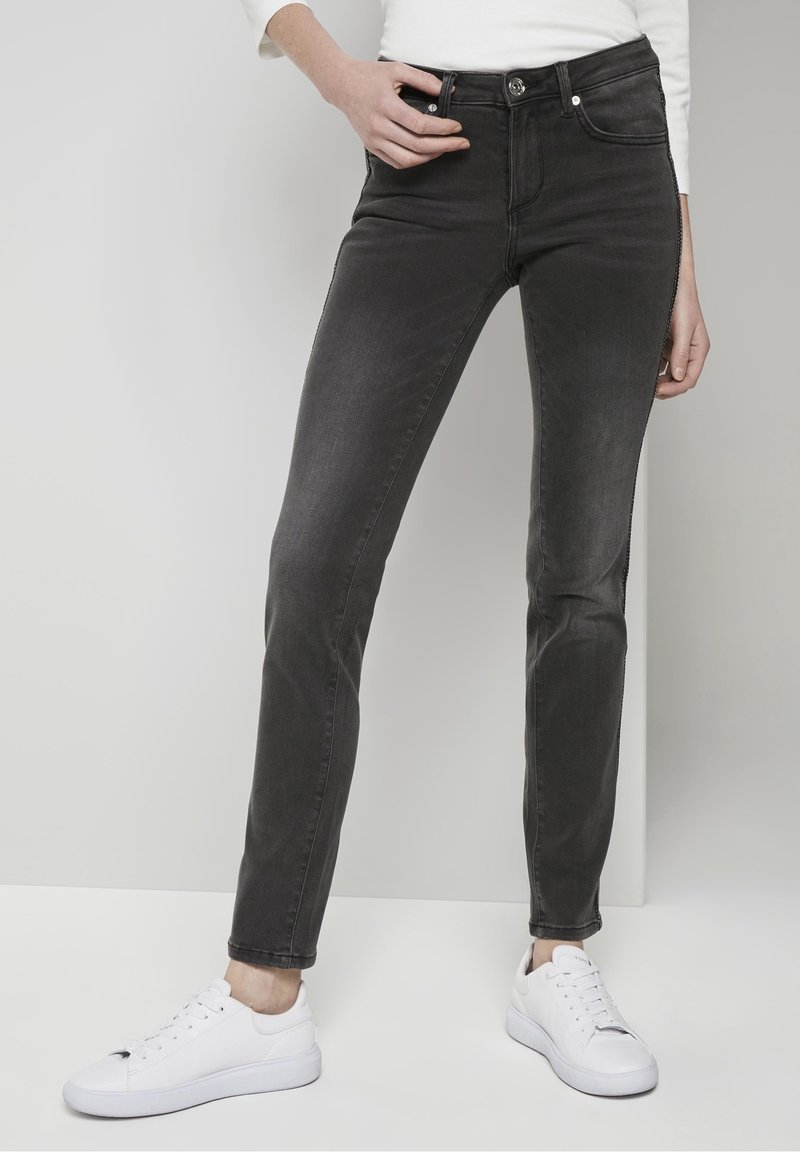 Jean noir slim-fit porté avec des baskets blanches et un haut blanc à manches longues, modèle debout devant un fond clair uni.