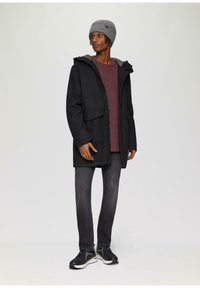 Schwarze Kapuzenjacke mit Fronttaschen, getragen über einem burgunderfarbenen Shirt, kombiniert mit grauen Jeans und schwarzen Sportschuhen. Graue Strickmütze auf dem Kopf.