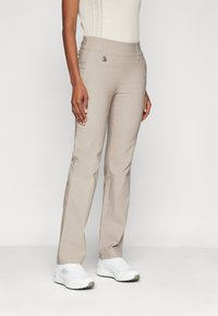 Pantalon beige avec une coupe droite, présentant une texture lisse, des poches avant et des détails de couture subtils, associé à des chaussures de sport blanches.