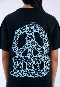 Camiseta de algodón negra con un gran símbolo de paz en azul claro formado por formas florales, rodeado de texto blanco.