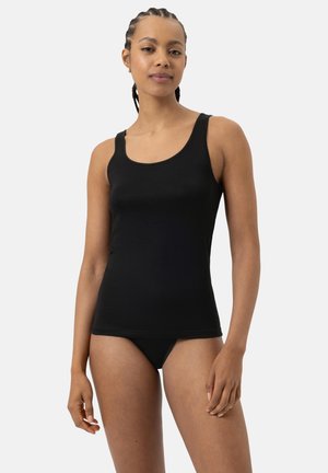 Donna con capelli intrecciati, che indossa una canotta nera e un coordinato di intimo nero, in piedi contro uno sfondo chiaro e semplice.