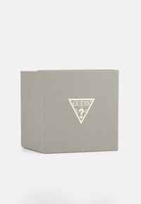 Šedá texturovaná dárková krabička s geometrickým tvarem, na které je zlaté logo ve tvaru trojúhelníku s nápisem "GUESS" a designem otazníku.