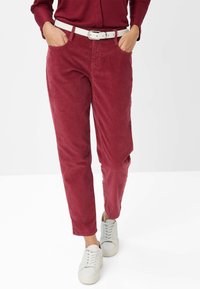 Pantaloni in corduroy cremisi con una vestibilità dritta, dotati di una cintura bianca e tasche laterali, abbinati a sneakers bianche su uno sfondo neutro.