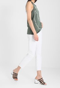 Zielona bezrękawnik z białym geometrycznym wzorem, zestawiony z białymi krótkimi spodniami i czarnymi sandałami w paski z obcasem w marmurkowy wzór.