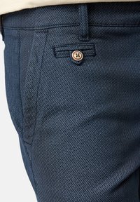 Gros plan sur un pantalon bleu foncé à motif montrant une poche latérale et une poche arrière boutonnée avec un tissu texturé.