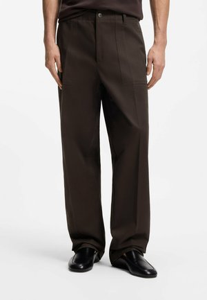 Man die een donkerbruine broek met wijd uitlopende pijpen en zwarte instapschoenen draagt, staand tegen een effen witte achtergrond.