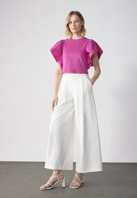 Magenta Kurzarm-Top mit Rüschensäumen, kombiniert mit weißen weiten Hosen und hellen Riemchen-Highheels auf einem grauen Boden.