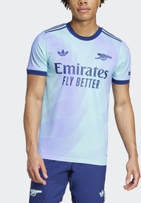 Lyseblå kortærmet trøje med bølgemønstre, marineblå accenter, "Emirates Fly Better" logo og Adidas-logo på brystet.