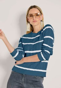 Frau mit großen beigefarbenen Sonnenbrillen, langen Perlenfransenohrringen, einem blau-weißen gestreiften Pullover aus Kabelstrick und grauen Jeans.