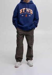 Person in einem blauen Hoodie mit dem Aufdruck "STWD", braunen Cargo-Hosen und weißen Sneakers, der mit den Händen in den Taschen vor einem einfarbigen Hintergrund steht.