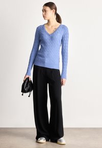 Gaiši zils džemperis ar rakstu, V-veida izgriezumu, garām piedurknēm, kopā ar melnām plaša piegriezuma biksēm. Modelis tur mazu melnu somiņu.