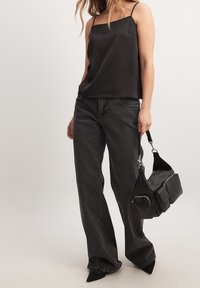Camisola nera in raso con spalline sottili, abbinata a jeans neri a vita alta e décolleté neri a punta, mentre tiene una borsa nera con texture.