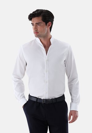 Camicia bianca a maniche lunghe con colletto alla mandarina, caratterizzata da una superficie liscia e chiusura a bottoni, indossata con pantaloni scuri e una cintura nera.