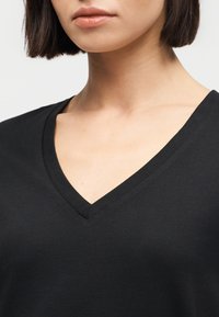 Γυναίκα με κοντά σκούρα μαλλιά που φοράει μαύρο μπλουζάκι με V λαιμόκοψη, εμφανίζοντας τον λαιμό, την κλείδα και το πάνω μέρος του στήθους, σε φωτεινό φόντο.