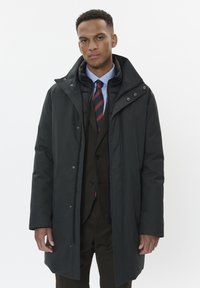 Melnas ārējās mēteļa ar augstu apkakli, kārtota virs tumšas uzvalka un svītrainas kaklasaite. Piedāvā rāvējslēdzēju un pogu aizdari, ar gludu tekstūru.