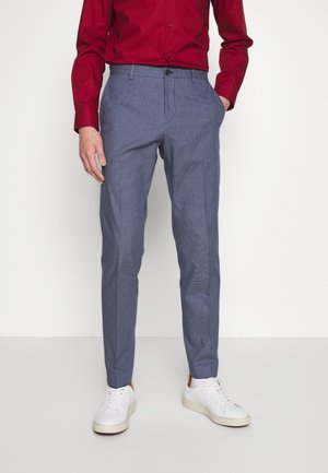 Homme portant un pantalon slim à carreaux bleus, des baskets blanches et une chemise rouge à manches longues, debout les mains dans les poches.