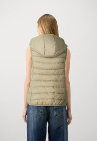 Puffväst i ljus olivgrön, ärmlös med huva. Har horisontell quiltad textur och dragkedjeförsedd fram. Bärs över blå jeans.