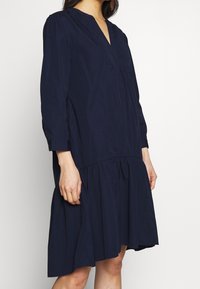 Mörkblå bomullsklänning med V-ringning, långa ärmar och en lagd, utsvängd fåll. Har subtil textur och rynkor under bysten.