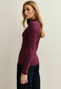 Långärmad, figurnära topp i djupröd ribbad tyg. Har en rund halsringning och kombineras med mörka, löst sittande jeans.