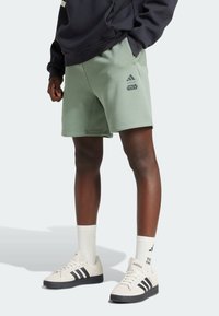 Osoba v světle zelených šortkách Adidas x Star Wars, černé mikině, bílých teniskách Adidas se černými pruhy a bílých kotníkových ponožkách.